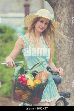 Attraktive blonde Frau mit Strohhut im Park stehen und posiert neben Fahrrad mit Korb voller Lebensmittel. Post-Geschäftsprozeß Stockfoto