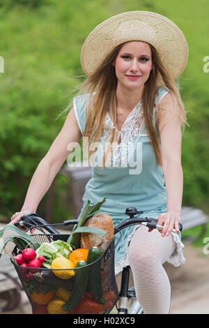 Attraktive blonde Frau mit Strohhut mit dem Fahrrad mit Korb voller Lebensmittel. Stockfoto