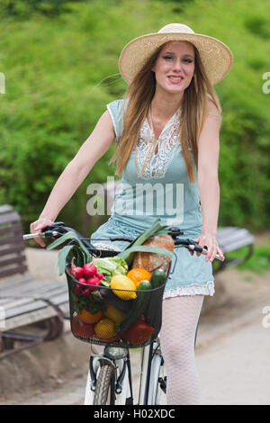 Attraktive blonde Frau mit Strohhut mit dem Fahrrad mit Korb voller Lebensmittel. Stockfoto