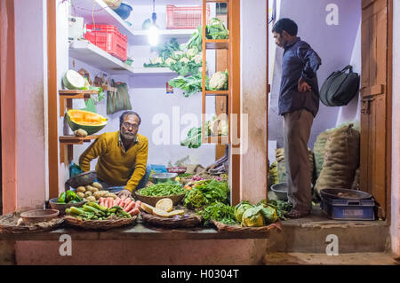 VARANASI, Indien - 19. Februar 2015: Lebensmittelhändler auf Boden sitzend mit Kunden stehen in der Tür kleine Gemüseladen auf Straße Ma Stockfoto