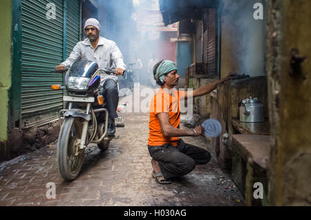 VARANASI, Indien - 20. Februar 2015: Straße Verkäufer macht Feuer für milchigen Tee in Kohle Ofen während Motorradfahrer vergeht. Stockfoto