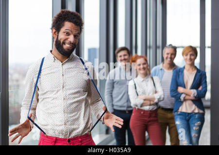 Unternehmensportrait der junge schwarze Hipster Geschäftsmann Dehnung Hosenträger, mit seinen Kollegen im Hintergrund. Stockfoto
