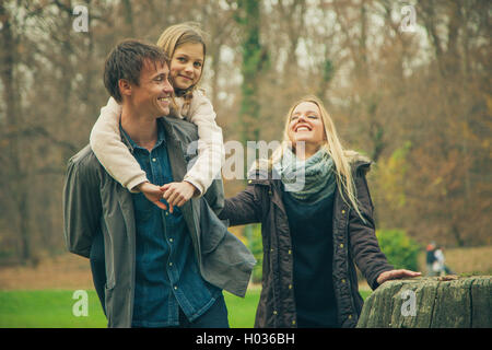 Tochter reitet Vater auf Huckepack mit Mutter neben ihnen im Park an einem Herbsttag. Stockfoto