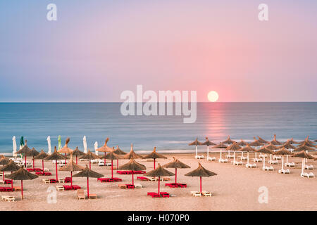 Klaren Himmel Sonnenaufgang über einen schwarzen Meer Sandstrand mit Stroh Sonnenschirme in Rumänien Stockfoto