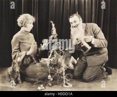Frau, die gerade Mann in falschen Bart Bewässerung Weihnachtsbaum Stockfoto