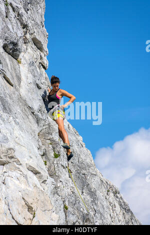 Gebräunte weibliche Kletterer in gelben Shorts auf steilen Felswand mit blauen Himmel und Wolken Stockfoto