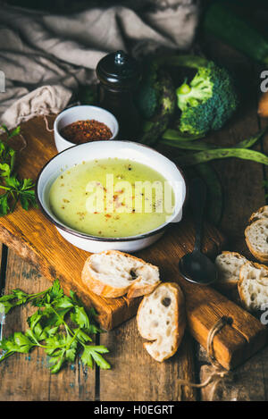 Frische hausgemachte Erbsen, Broccoli, Zucchini Creme Suppe in weiße Schüssel mit frischem Baguette Scheiben und Petersilie auf Holzbrett über Rost Stockfoto