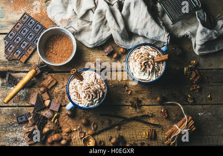 Heiße Schokolade mit Sahne, Nüssen und Zimt in Emaille Becher mit Zutaten aller auf rustikalen hölzernen Hintergrund, Ansicht von oben Stockfoto