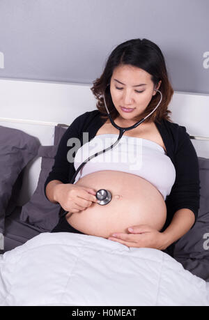 Schwangere Frau mit einem Stethoskop auf dem Bett auf den Bauch hören Stockfoto