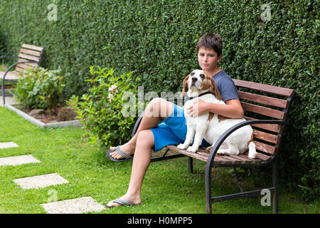 Glückliche junge und sein Hund Beagle sitzend auf Bank im Frühlingsgarten. Junge umarmt seinen Hund Stockfoto