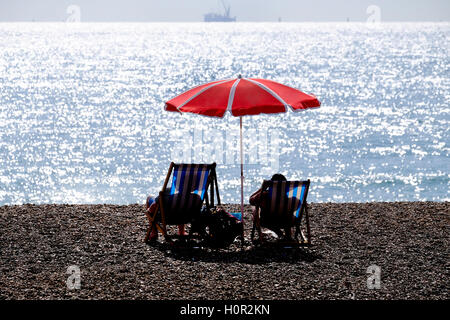 die Silhouetten von zwei Menschen sitzen in Liegestühlen am Kieselstrand unter einem roten Regenschirm, Blick auf das Meer Stockfoto