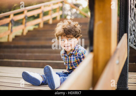 Porträt von kleinen niedlichen Kind sitzt auf Holztreppen im Stadtpark. Junge sieht in die Kamera durch hölzerne Geländer der Treppe Stockfoto