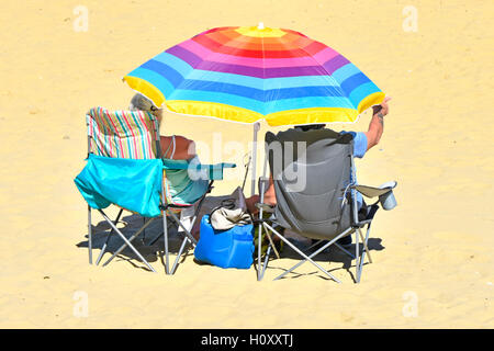 Seniorenpaar unter buntem Sonnenschirm entspannender Lifestyle Klappstühle Sandstrand Britischer Badeurlaub im Schatten von heißer Sommersonne UK Stockfoto