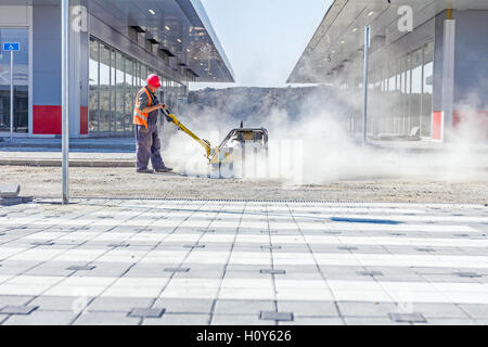 Arbeitnehmer arbeitet mit Vibration Platte Müllpresse Maschine vor der Baustelle Gehsteig. Staub ist herumfliegen. Stockfoto