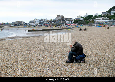 Ein Obdachloser Rucksack trinken Kaffee sitzen am Strand, Blick auf das Meer bei Lyme Regis, Dorset, England UK KATHY DEWITT Stockfoto