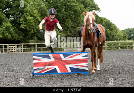 Ein junges Pony Reiter Springen einen niedrigen Zaun A Chestnut Pony schaut zu, wie der Fahrer springt einen niedrigen Zaun zeigt die UK-Anschluß-Markierungsfahne Stockfoto