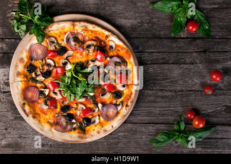 Leckere frische Pizza mit Zwiebeln, schwarzen Oliven, Paprika, Champignons und Rucola auf dem hölzernen Hintergrund. Ansicht von oben. Stockfoto