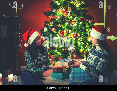 Junge weibliche Freunde in Hüte santa claus in der Nähe einen Weihnachtsbaum in der dekorierten Zimmer Geschenke zu jedem anderen gibt Stockfoto