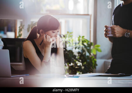 Schuss von junge Frau sitzt an ihrem Schreibtisch und Gespräch mit männlichen Kollegen stehen. Mitarbeiter arbeiten gemeinsam an Neugeschäft Stockfoto