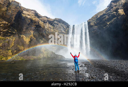 Wanderer vor der Skógafoss Wasserfall mit ausgestreckten Armen, Regenbogen, Skogar, Region Süd, Island Stockfoto