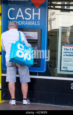 Ein senior Mann in Shorts und Trainer mit einer Co-Op-Tasche zieht Geld aus einer Nationwide Building Society Geldautomaten Stockfoto