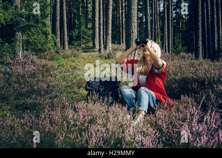 Junge aktive Frau Tourist sitzt in einer Lichtung im Wald und auf der Suche durch ein Fernglas. Gesunden, aktiven Lebensstil-Konzept. Stockfoto