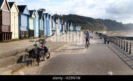 Für immer reife junge - Dame Wellen Abschied von ihren Freunden, wie sie ihre entlang der Strandpromenade, gegenüber bunten Hütten Radtouren. Stockfoto