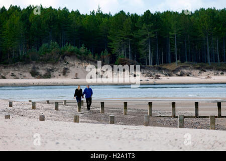 Ein paar Der sroll entlang der Strand in Findhorn Bay, Schottland mit der dicken Vertrauen Wald in der Ferne, Großbritannien Stockfoto