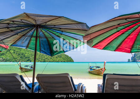 Blick auf long-Tail-Boote und Meer durch Liegestühle und Sonnenschirme am Strand von Ko Phi Phi Don, Thailand Stockfoto