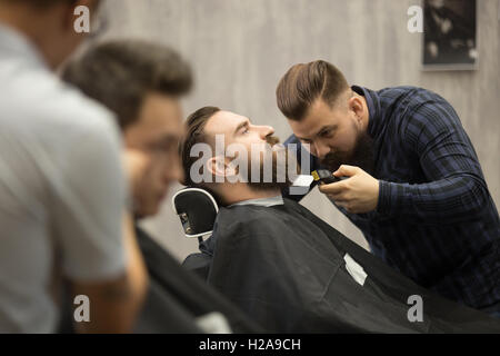 Innenaufnahme des Arbeitsprozesses im Barbershop. Seitenansicht der hübschen jungen Mann immer trendy Bart Haarschnitt in modernen barbersho Stockfoto