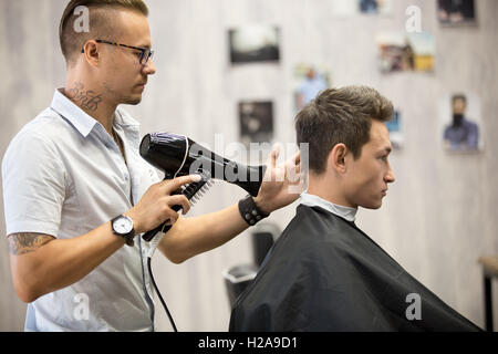 Arbeitsprozess im modernen Friseursalon. Gut aussehend Friseur servieren Kunden, Haar für männliche Kunden mit Föhn-styling. Seite Stockfoto