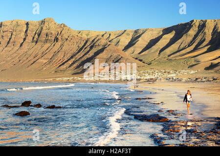 Junge Frau zu Fuß am Strand Playa La Caleta de Famara mit Klippen von Risco de Famara. Lanzarote, Kanarische Inseln Stockfoto
