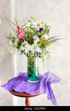 Farbenfrohe Blumenarrangements mit Lilien, Rosen und Grün in einer Glasvase auf einem Tisch, mit violettem Stoff an einer hellen Wand. Stockfoto