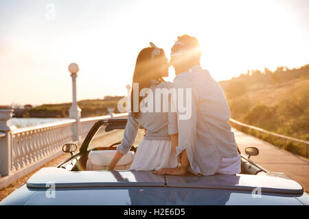 Glückliches junges Paar sitzt und küssen im Cabriolet auf Sonnenuntergang Stockfoto