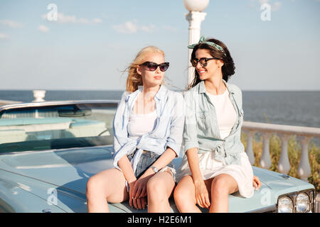 Zwei lächelnde attraktive junge Frauen sitzen auf dem Auto im Sommer Stockfoto