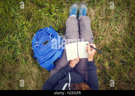 Frau mit blauen Rucksack auf dem Rasen sitzen und schreiben in leere Arbeitsmappe. Über Kopf warm getönten Farbansicht Stockfoto