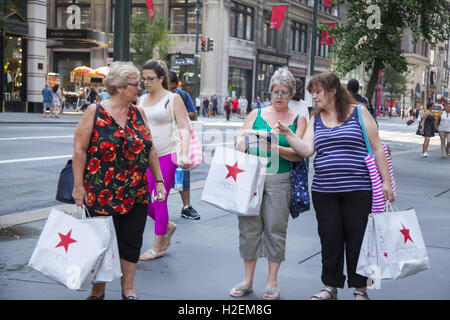 Drei Frauen, die alle mit Macy's Shopping Taschen auf der 5th Avenue in Midtown Manhattan. New York City Consulting eine touristische Broschüre Weitere Informationen wohin man als nächstes gehen soll. Stockfoto