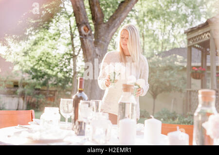 blonde Frau einrichten einen Tisch in einem Garten, Kerzen und Vasen mit rosa Rosen. Stockfoto