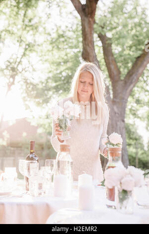 blonde Frau einrichten einen Tisch in einem Garten, Kerzen und Vasen mit rosa Rosen. Stockfoto