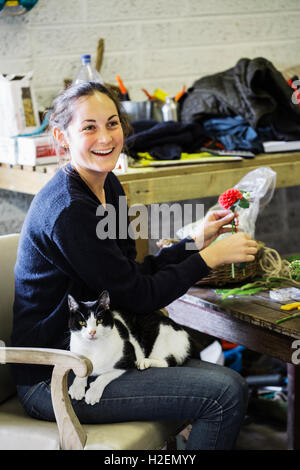 Eine Frau in einer Scheune Blume arbeiten über eine Vereinbarung mit einer Katze auf dem Schoß sitzen. Stockfoto