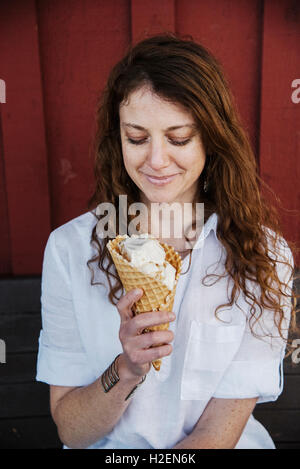 Frau mit Lon g braunen Haaren sitzen auf einer Bank, Eis essen. Stockfoto