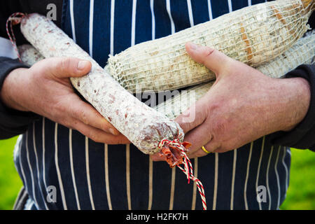 Nahaufnahme eines Metzgers trägt eine gestreifte blaue Schürze, im freien stehend, hält eine Auswahl von Salamis. Stockfoto