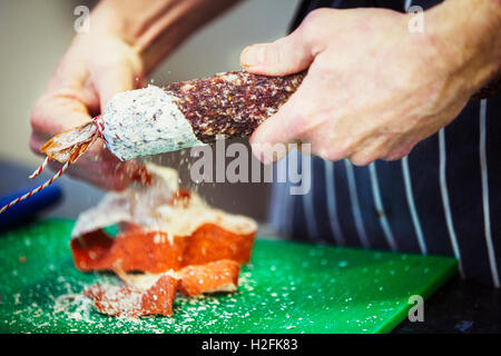 Nahaufnahme eines Metzgers trägt eine gestreifte blaue Schürze, unter der Haut eine Salami. Stockfoto