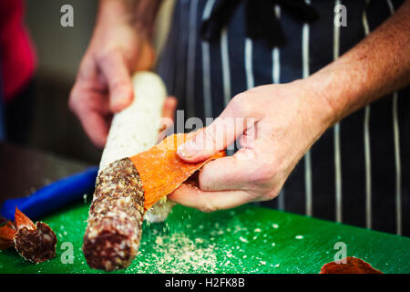 Nahaufnahme eines Metzgers trägt eine gestreifte blaue Schürze, unter der Haut eine Salami. Stockfoto