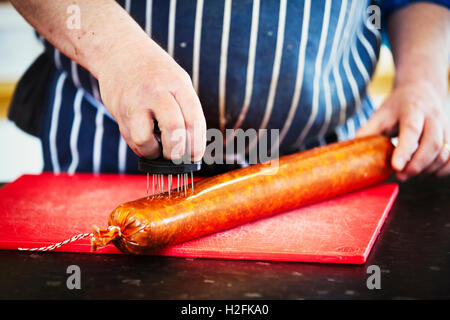 Nahaufnahme eines Metzgers trägt eine gestreifte blaue Schürze stechen Löcher in das Gehäuse einer Chorizo-Wurst. Stockfoto