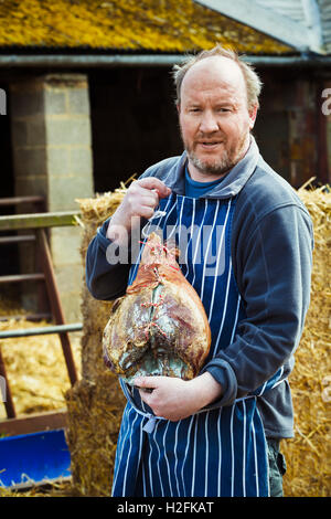 Metzger trägt eine gestreifte blaue Schürze, im Freien stehen, halten einen großen Schinken. Stockfoto