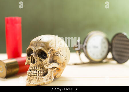 Menschlicher Schädel ruht auf einem Holzfußboden und einer roten Schrotflinte Shell mit antiken Uhren im Hintergrund. Stockfoto
