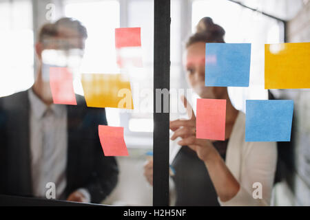 Zwei Kollegen zusammen am Projekt arbeiten. Business-Frau zeigt auf Haftnotiz an männlichen Kollegen auf Glaswand in o Stockfoto