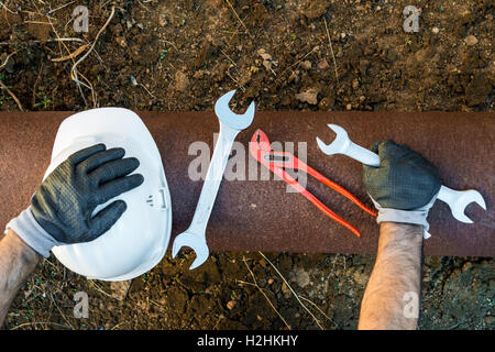 Hände mit Arbeitshandschuhen hält einen Schutzhelm, einen Schraubenschlüssel und einen roten Schlüssel Rohr klemmt Stockfoto