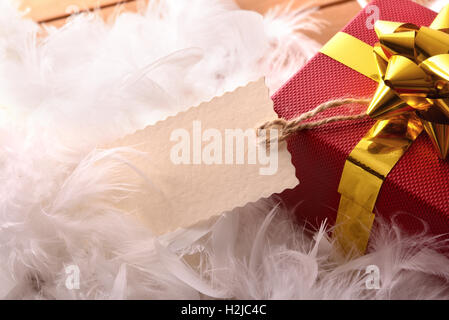 Roten Geschenkbox mit goldenen Band und Tag hängen Closeup auf weißen Federn und weißem Hintergrund. Erhöhten Draufsicht. Stockfoto